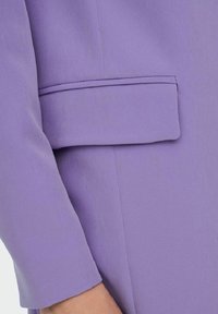 Lila bleiseri tasaisella tekstuurilla; siinä on läpällinen tasku vasemmassa hihassa ja muotoiltu design, jossa on 3/4-pituiset hihat.