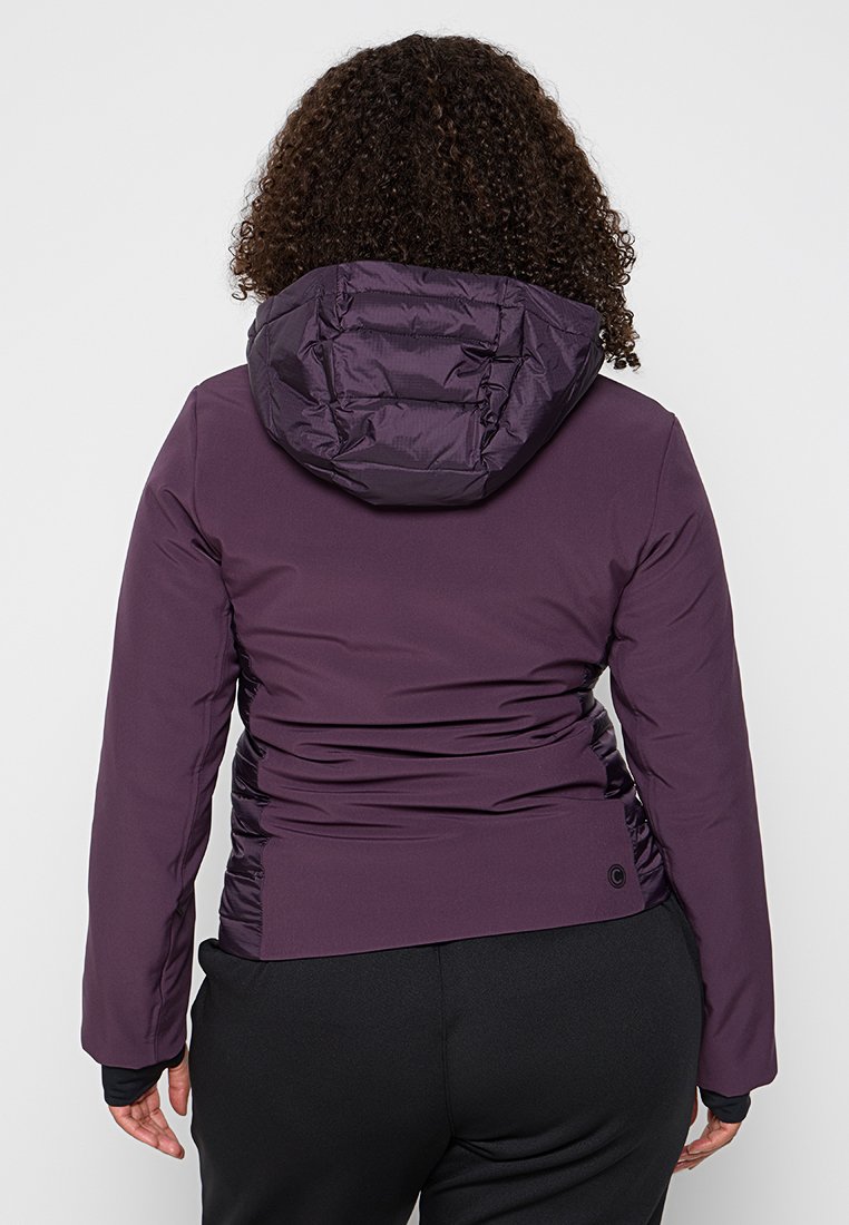 Giacca isolante viola con design aderente, cappuccio voluminoso e dettagli arricciati sui lati. Il tessuto leggero e i polsini elasticizzati aumentano il comfort.
