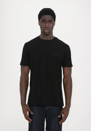 T-shirt en coton noir à col rond avec des manches courtes et un petit logo ton sur ton sur la poitrine. Le tissu est lisse et léger. Assorti à un jean bleu foncé.