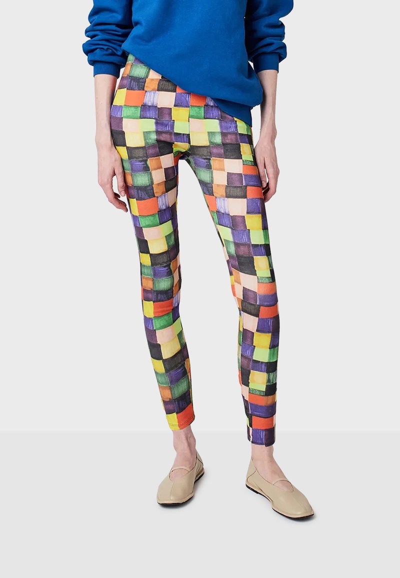 Persoon in kleurrijke geruite leggings met oranje, groen, paars en gele vierkanten, beige flats en een oversized blauwe sweatshirt.