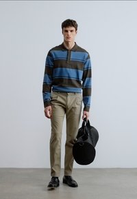 Πόλο με ριγέ μακριά μανίκια σε μπλε και καφέ με γιακά· ανοιχτό μπεζ παντελόνι· κρατώντας μια μαύρη στρογγυλή τσάντα duffel· στέκεται σε ουδέτερη επιφάνεια.