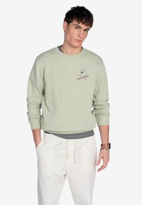 Sudadera verde claro con puños acanalados, que presenta un diseño de logo en el pecho. Combinada con pantalones blancos con cordón. Textura suave.