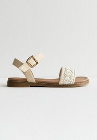 Sandale beige avec un tissu blanc tissé à l'avant, sangle de cheville ajustable avec boucle, semelle rembourrée marron. Design plat, style décontracté.