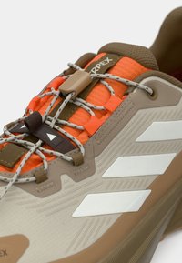 Μπεζ και πορτοκαλί πεζοπορικό παπούτσι με υφασμάτινη επάνω επιφάνεια με υφή, ελαστικά κορδόνια και καφέ λεπτομέρειες. Διαθέτει τρεις λευκές ρίγες στο πλάι.