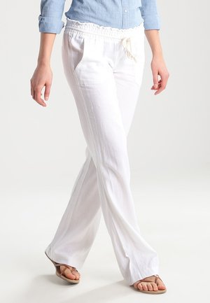 Mujer vestida con pantalones blancos anchos con cordón, camisa azul claro con mangas remangadas y sandalias beige, caminando con las piernas cruzadas.