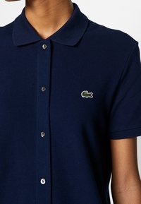 Polo bleu marine en tissu texturé, avec col, boutons argentés et petit logo crocodile vert sur la poitrine.