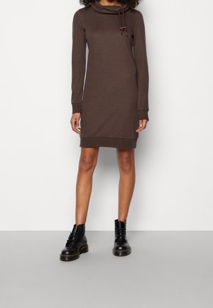 Femme portant une robe à capuche marron à manches longues, arrivant aux genoux, avec des bottines noires à lacets, debout sur un sol gris contre un mur blanc.