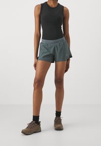 Top negro sin mangas y pantalones cortos color verde azulado hechos de tela ligera. Detalles destacados incluyen una cintura ancha y un logo en los pantalones cortos. Zapatos de senderismo marrones.