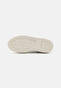 Semelle de chaussure en caoutchouc blanc avec motifs antidérapants texturés et logo de la marque sur un fond gris clair uni.