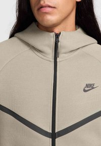Beige huppari, joka on valmistettu teksturoidusta kankaasta, edessä vetoketju mustilla kiinnikkeillä ja Nike-logo painettuna vasempaan rinta-alueeseen.