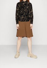 Svart blus med guldblommönster, brun A-linjeformad kjol och beige tjocka loafers. Slät tyg och väldefinierad passform synlig.