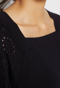 Svart stickad tröja med fyrkantig halsringning, som har texturerade sidopaneler och små hål för ventilation. Slät, åtsittande tyg.