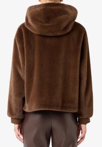 Persona che indossa un giubbotto di pelliccia pelosa marrone scuro con cappuccio, abbinato a pantaloni di pelle marrone scuro, mostrata di spalle.