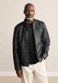 Schwarze Lederjacke mit hohem Kragen, Frontreißverschluss und zwei Seitentaschen. Über einem schwarzen Shirt getragen, kombiniert mit hellfarbigen Hosen.