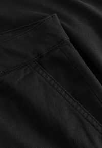 Fekete szövet texturált felülettel, átlós varrással és a bal felső sarokban rendezetten varrt zsebbel.