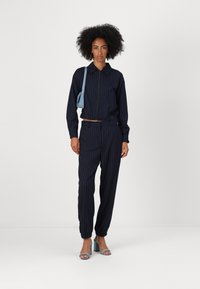 Tuta a righe blu scuro composto da una giacca con zip, colletto e polsini elasticizzati, abbinato a pantaloni affusolati e sandali con tacco azzurri chiari.