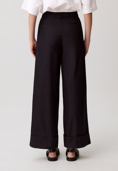 Pantaloni neri a gamba larga con una texture liscia, caratterizzati da una vita alta, risvolti arrotolati e due tasche posteriori.