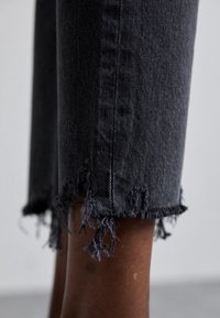 Vaqueros de mezclilla negra con corte recto, con un dobladillo deshilachado y un detalle textural. La tela tiene un acabado suave y texturado.