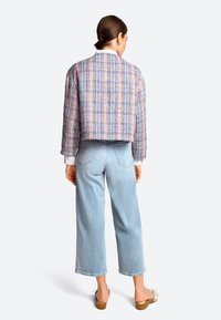 Gekürzte, mehrfarbige Tweedjacke mit blauen, roten und weißen Mustern; kombiniert mit hellblauen, weiten Jeans und braunen Sandalen.