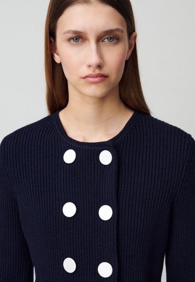 Cardigan en maille côtelée marine avec de grands boutons blancs, col rond et tissu texturé. Gros plan sur le détail de la partie supérieure du vêtement.