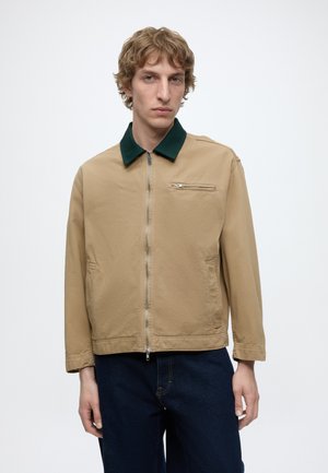 Jeune homme portant une veste beige zippée avec un col vert foncé, une poche zippée avant, et un jean bleu foncé, debout devant un fond blanc uni.