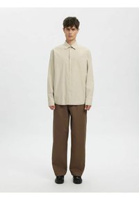 Langermet, lett rutete skjorte i beige med knappestolpe og krage, kombinert med løse brune bukser. Svarte sko. Enkel design.