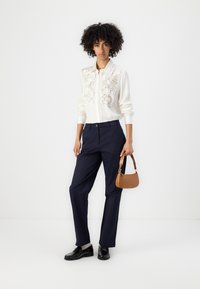 Weißes Blusenshirt mit floralem Muster auf der Vorderseite, marineblaue Hose, schwarze Schuhe und eine kleine braune Handtasche. Einfaches und modernes Design.