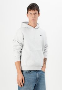 Fleece cinza claro com capuz e um bolso tipo canguru, apresentando um pequeno logótipo preto "OBEY" no peito e punhos canelados. Jeans de ganga usados por baixo.