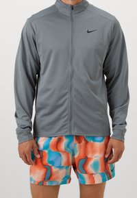 Grå zip-up jakke med en glat tekstur, lange ærmer og et diskret Nike-logo. Sammen med farverige shorts med et livligt mønster.