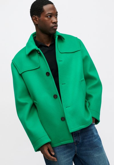 Chaqueta de lana verde con un amplio cuello, cierre de botones y bolsillos laterales. La tela tiene una textura suave y se combina con unos jeans de mezclilla oscuros.