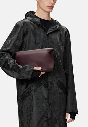 Una persona joven lleva un impermeable negro brillante con capucha y sostiene un estuche rectangular mate de color marrón oscuro cerca del pecho.
