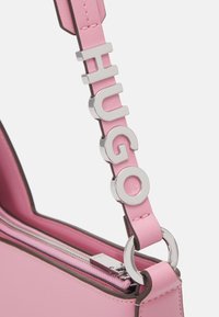 Anse de sac en cuir rose avec des lettres métalliques argentées formant le mot HUGO, attachée à un anneau argenté et un curseur de fermeture éclair rose.