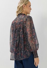 Transparente schwarze Bluse mit rotem Blumenmuster, gepunktetem Design, hohem Kragen und gerafften Ärmelabschlüssen. Lange Ballonärmel sorgen für eine fließende Silhouette.
