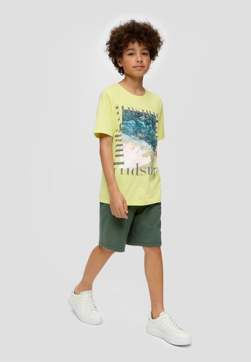 Geel t-shirt met korte mouwen en grafische print, gecombineerd met groene shorts en witte sneakers. De stof lijkt zacht en casual.