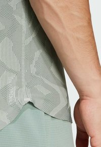 Ljusgrön atletisk t-shirt med texturerad mönster, korta ärmar och en lätt nedre fåll bak, tillverkad av andningsbart material.
