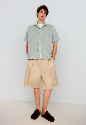 Ung mand iført en kortærmet, stribet skjorte med knapper, beige shorts til knæet og mørke slip-on sko, stående mod en ensfarvet væg.