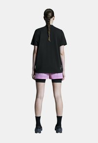 Schwarzes T-Shirt mit kurzen Ärmeln, kombiniert mit rosa Shorts. Das Modell trägt schwarze Socken und schwarze Sportschuhe. Einfaches Design, keine auffälligen Muster.
