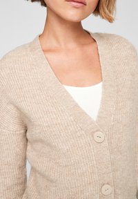 Beige gebreide cardigan met een gemêleerd patroon, v-hals en twee knoopsluitingen. Zacht, middelzwaar stof. Daaronder een witte top.
