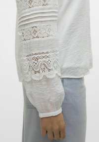 Camicia bianca a maniche lunghe con tessuto testurizzato, caratterizzata da dettagli in pizzo, pieghe sulle maniche e polsini aderenti, abbinata a jeans chiari.