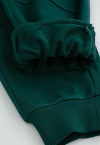 Zielona koszulka z materiału z ściągaczem. Ściągacz ma teksturę marszczoną, podczas gdy korpus jest gładki z delikatnymi detalami stębnowania.