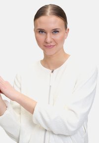 Weiße Zip-Jacke aus strukturiertem Rippmaterial mit Pailletten. Sie besitzt einen runden Ausschnitt und elastische Bündchen. Das Modell posiert mit einer erhobenen Hand.