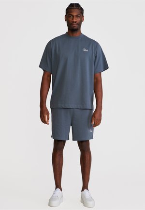 Mann som står vendt framover, iført matchende mørkegrå oversized t-skjorte og shorts med hvite joggesko på ensfarget bakgrunn.