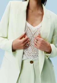 Femme portant un blazer crème et un pantalon sur un haut en dentelle crochet blanc, ajustant sa veste avec ses mains ornées de bagues en argent et en perles.