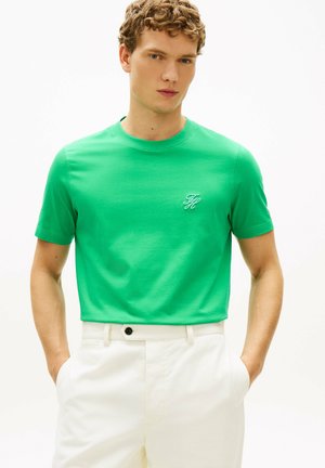 Giovane uomo con camicia verde brillante a maniche corte con piccolo logo bianco e pantaloni bianchi, mani in tasca, sfondo neutro.