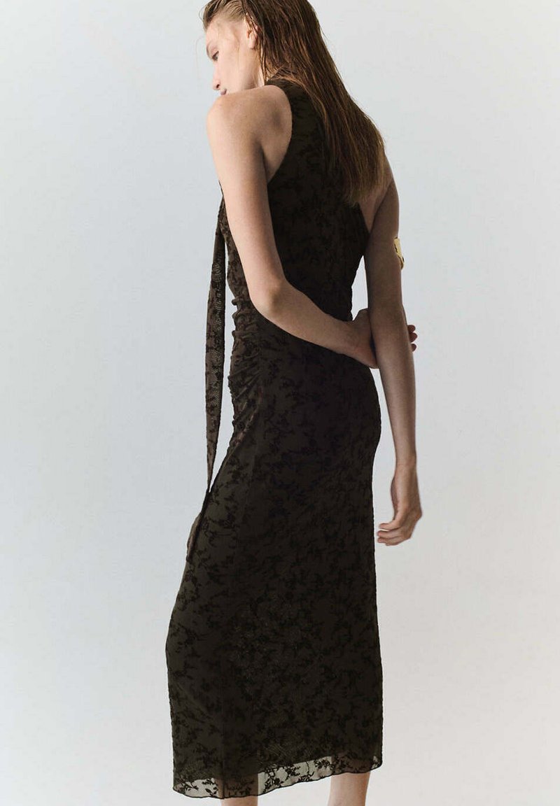 Femme aux longs cheveux portant une robe en dentelle sombre sans manches, longue jusqu'aux chevilles, debout devant un fond clair uni.