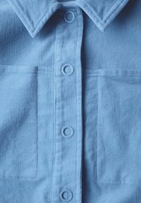 Chemise en velours côtelé bleu clair avec col pointu, boutons de fermeture et deux poches avant. Texture lisse avec un motif côtelé.