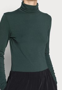 Frau in einem dunkelgrünen, figurbetonten Langarmrollkragenpullover und einer lockeren schwarzen Hose mit elastischem Bund, stehend vor einem einfarbigen Hintergrund.