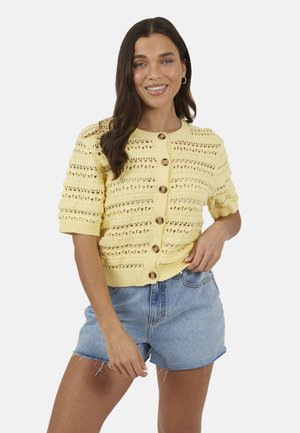 Donna sorridente con lunghi capelli scuri che indossa un cardigan giallo a maniche corte lavorato all'uncinetto con bottoni e pantaloncini di jeans azzurri.