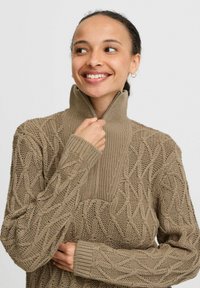 Beige gebreide trui met een hoge kraag en ritssluiting, voorzien van een structuur kabelpatroon en geribbelde accenten bij de hals en mouwen.