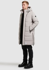 Cappotto imbottito grigio chiaro con cappuccio, caratterizzato da un design ampio, tasche laterali con zip e un'etichetta con il marchio vicino all'orlo. Indossato sopra una maglietta nera e jeans.